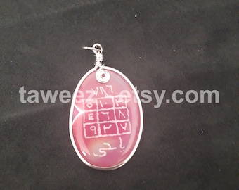 May include: A pink oval pendant with a silver wire wrap and a silver bail. The pendant features a white inscription of a square with Hebrew letters inside. The pendant is on a black background.