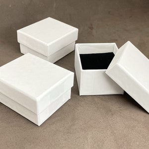 Puede incluir: Tres cajas de cart&oacute;n blancas con tapas. Dos cajas est&aacute;n cerradas y una caja est&aacute; abierta con un interior de terciopelo negro.