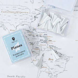 Peut inclure: Des punaises en forme d'avion en papier blanc dans une boîte transparente, avec une carte indiquant "Planes 10 paper plane push pins". Les articles sont disposés sur une carte du monde.
