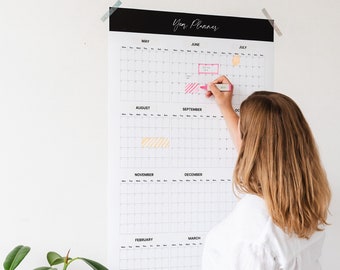May include: A white wall calendar with black headings for each month. The calendar is labeled "Year Planner" and includes the months of May through March. A person is using a pink highlighter to mark a date on the calendar.