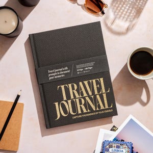 以下が含まれることがあります： 「TRAVEL JOURNAL」と「CAPTURE THE ESSENCE OF YOUR TRAVELS」という金色の文字が書かれた黒い旅行日記。日記には、「Travel journal with prompts to document your memories」と「16 Trips / 205 Pages」と書かれた黒いバンドが巻かれている。