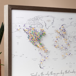 Może przedstawiać: Biała mapa świata w drewnianej ramie. Mapa jest pokryta kolorowymi szpilkami, oznaczającymi miejsca na całym świecie. Tekst "Travel is the only thing you buy that makes you richer" jest wydrukowany na dole mapy.