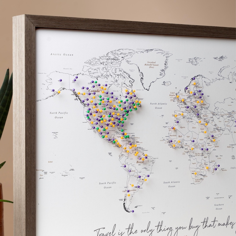Może przedstawiać: Biała mapa świata w drewnianej ramie. Mapa jest pokryta kolorowymi szpilkami, oznaczającymi miejsca na całym świecie. Tekst "Travel is the only thing you buy that makes you richer" jest wydrukowany na dole mapy.