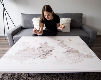 May include: A person sits on a gray couch and places a small, round, gold pin on a large world map printed on a white surface. The map is laid out on a white coffee table with black legs.