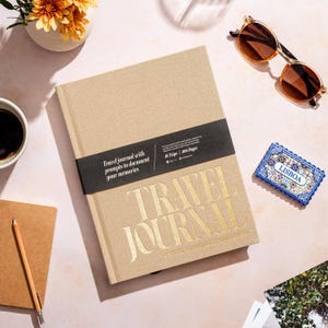Puede incluir: Un diario de viaje beige con letras doradas y la inscripción "TRAVEL JOURNAL". El diario incluye indicaciones para documentar recuerdos y tiene una banda negra con texto. Otros elementos incluyen gafas de sol, una taza de café y una baldosa decorativa.