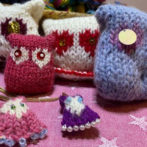 May include: Handmade knitted owl figurines in various colors, including pink, white, and blue. Some owls have decorative sequins and beaded accents. The owls are arranged on a pink patterned surface, suggesting a craft or home decor theme.