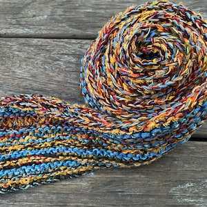 May include: A hand-knitted scarf featuring a variegated yarn blend of mustard yellow, deep burgundy, and sky blue. The accessory is coiled into a circular shape, showcasing a consistent garter stitch pattern throughout the entire length of the fabric.