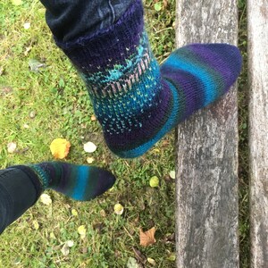 May include: A pair of colorful hand-knitted socks with a blue, green, and purple color scheme. The socks are worn on a person's feet and are resting on a wooden surface.