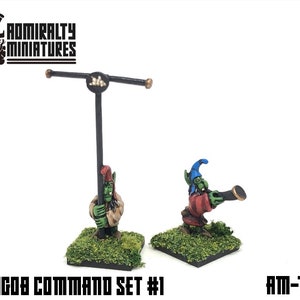 Puede incluir: Dos miniaturas de duendes verdes, una sosteniendo un asta de bandera y la otra un telescopio, sobre una base verde. Las miniaturas forman parte de un set de mando para un juego de mesa. El texto "T.J.W.8608 COMMAND SET #1" y "AM-T29" es visible en la imagen.