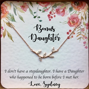 Puede incluir: Una tarjeta con un diseño floral y el texto "Bonus Daughter" y "No tengo una hijastra. Tengo una hija que nació antes de que la conociera. Con amor, Sydney". Un collar de oro rosa con un diseño de rama y hojas está encima de la tarjeta.