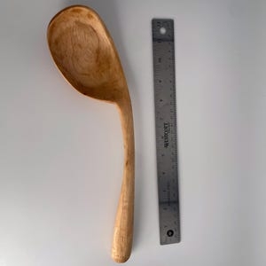 May include: A handcrafted wooden spoon with a curved handle and a large, rounded bowl. The spoon is made of light-coloured wood and measures approximately 30 cm in length. A metal ruler is next to the spoon.