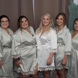 May include: Five individuals wearing matching satin robes. Four people wear light sage green robes with white lace trim on the sleeves and hem, while the central person wears a white satin robe with matching white lace trim on the sleeves and hem.