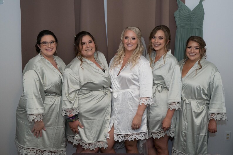 May include: Five individuals wearing matching satin robes. Four people wear light sage green robes with white lace trim on the sleeves and hem, while the central person wears a white satin robe with matching white lace trim on the sleeves and hem.