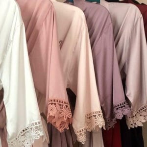 May include: A row of satin robes hanging on a rack, featuring wide sleeves with intricate lace trim at the cuffs. The robes come in various colors including white, blush pink, mauve, burgundy, and navy, with the word Bride printed on the white robe.