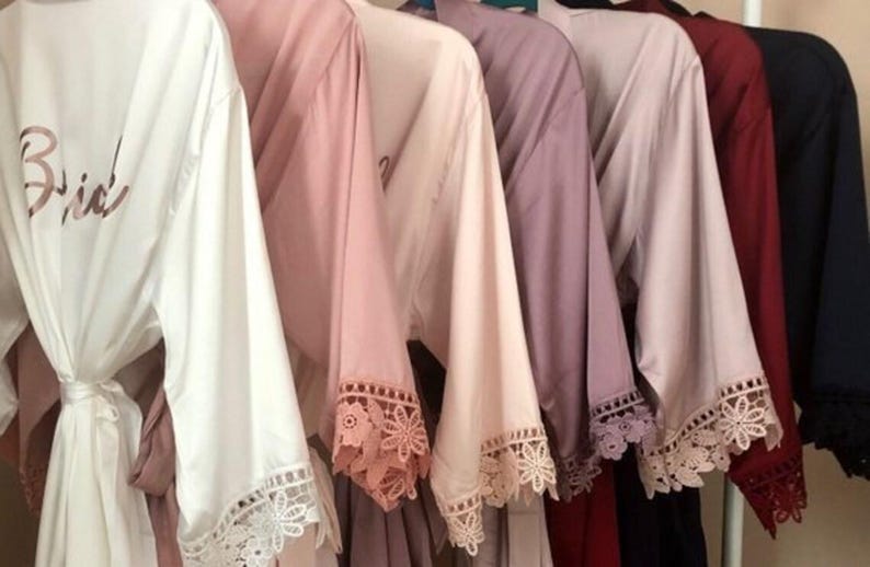 May include: A row of satin robes hanging on a rack, featuring wide sleeves with intricate lace trim at the cuffs. The robes come in various colors including white, blush pink, mauve, burgundy, and navy, with the word Bride printed on the white robe.