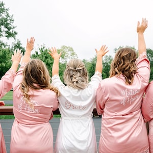 May include: Five women wearing pink satin robes with white embroidery. The robes say "Bride", "Matron of Honor", "Maid of Honor", and "Bridesmaid".