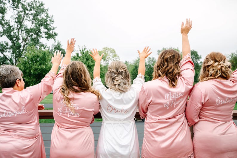 May include: Five women wearing pink satin robes with white embroidery. The robes say "Bride", "Matron of Honor", "Maid of Honor", and "Bridesmaid".