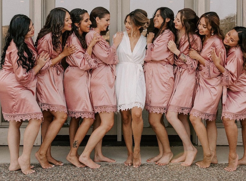 May include: A group of nine individuals wearing matching satin robes. Eight individuals wear dusty rose pink robes with lace trim on the sleeves and hem, while one individual in the center wears a white robe with matching lace trim details.