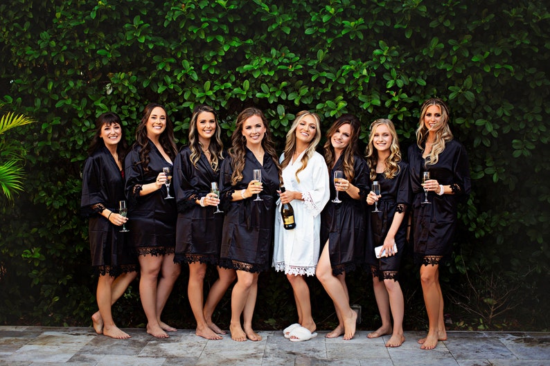 May include: A group of eight individuals wearing matching robes, seven in black and one in white, each featuring lace trim on the sleeves and hem. The individuals are standing in a row, with several holding champagne flutes and one holding a bottle.