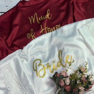 May include: Two satin robes with lace trim. One is white with the word "Bride" in gold lettering. The other is burgundy with the words "Maid of Honor" in gold lettering.