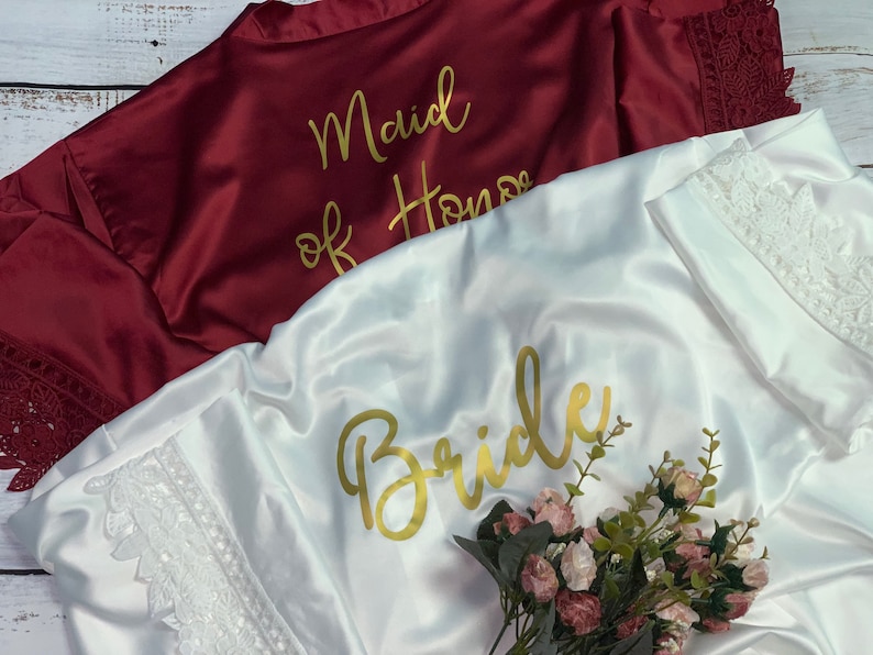 May include: Two satin robes with lace trim. One is white with the word "Bride" in gold lettering. The other is burgundy with the words "Maid of Honor" in gold lettering.