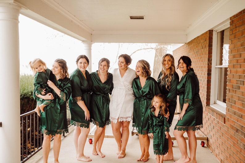 May include: A group of women and girls wearing green satin robes with lace trim. The women are smiling and looking at the camera. The girls are looking at the camera. The women are standing on a porch with a brick wall in the background.