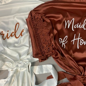 May include: Two satin robes with lace trim. One robe is white with the word "Bride" in brown lettering. The other robe is brown with the words "Maid of Honor" in white lettering.
