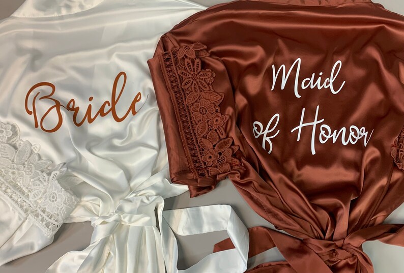 May include: Two satin robes with lace trim. One robe is white with the word "Bride" in brown lettering. The other robe is brown with the words "Maid of Honor" in white lettering.