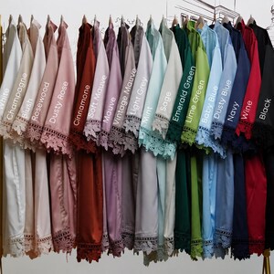 May include: A collection of satin robes hanging on a gold rack, each featuring lace trim on the sleeves and hem. The robes are labeled with colors: White, Champagne, Blush, Rosewood, Dusty Rose, Cinnamone, Soft Mauve, Mauve, Vintage Mauve, Light Grey, Mint, Sage, Emerald Green, Lime Green, Baby Blue, Dusty Blue, Navy, Wine, and Black.