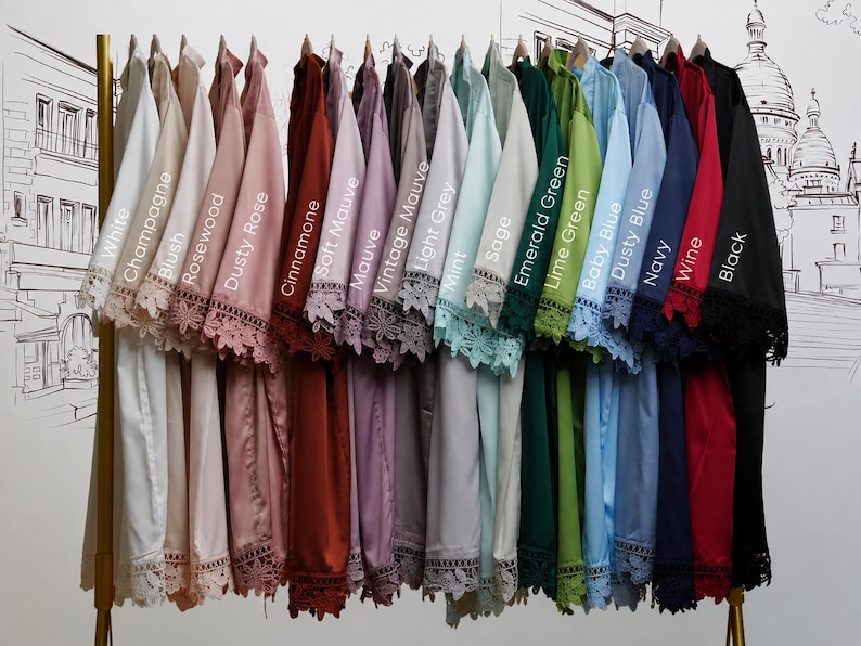 May include: A collection of satin robes hanging on a gold rack, each featuring lace trim on the sleeves and hem. The robes are labeled with colors: White, Champagne, Blush, Rosewood, Dusty Rose, Cinnamone, Soft Mauve, Mauve, Vintage Mauve, Light Grey, Mint, Sage, Emerald Green, Lime Green, Baby Blue, Dusty Blue, Navy, Wine, and Black.