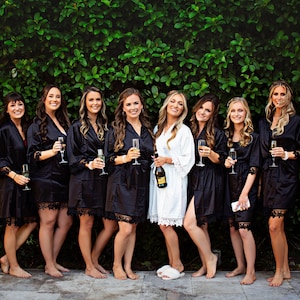 May include: A group of eight people wearing black and white robes stand in front of a green wall. They are holding glasses of champagne and smiling.
