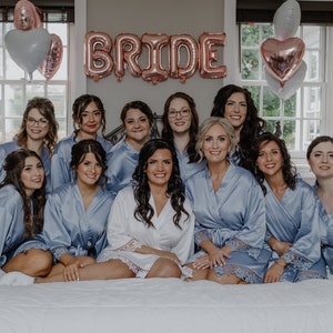 May include: A group of women in matching light blue satin robes with lace trim, posing for a photo. The bride is in the center wearing a white satin robe with lace trim. The word "BRIDE" is spelled out in large, rose gold balloons above them.