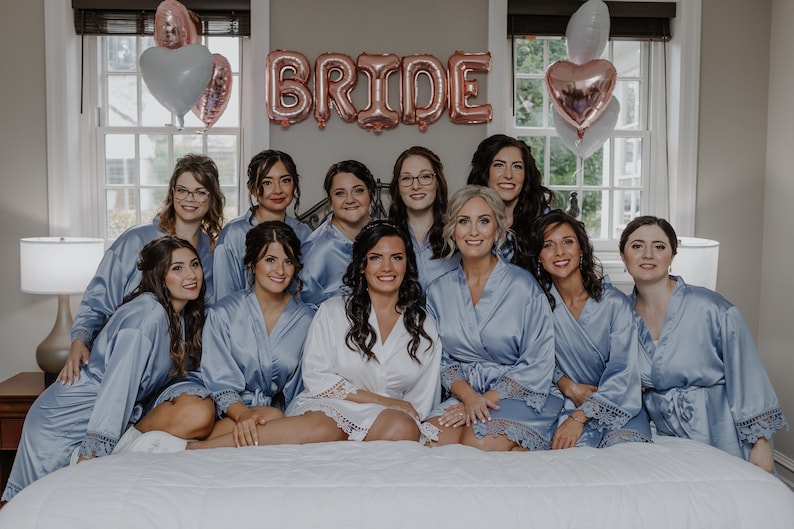 May include: A group of women in matching light blue satin robes with lace trim, posing for a photo. The bride is in the center wearing a white satin robe with lace trim. The word "BRIDE" is spelled out in large, rose gold balloons above them.