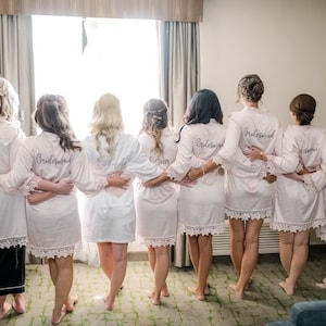 May include: A group of seven individuals standing in a row, wearing matching light pink robes with white lace trim on the sleeves and hems. Each robe features text embroidered on the back, including the words Bride, Maid of Honor, and Bridesmaid.