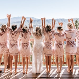 May include: Eight people wearing pink satin robes with lace trim stand on a wooden deck with their arms raised in the air. They are looking out at a mountain range in the distance.