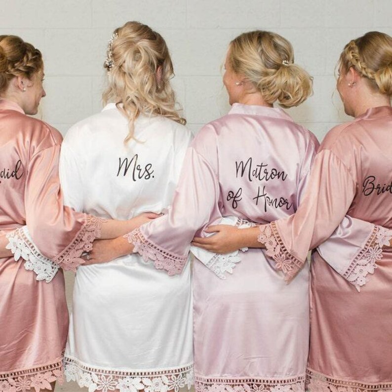 May include: Four satin robes with lace trim on the sleeves and hem. The robes are in shades of dusty rose and white. The white robe features the text 'Mrs.' on the back, while the dusty rose robe features the text 'Matron of Honor' on the back.