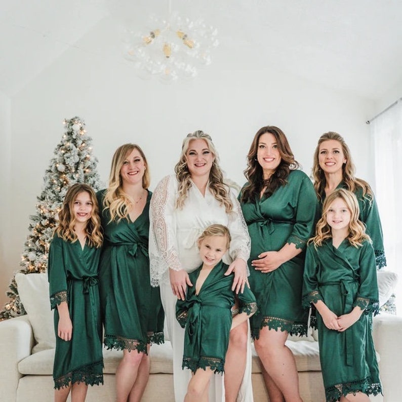 May include: A group of people wearing green satin robes with lace trim. The robes are tied at the waist and have long sleeves. The people are standing in front of a white wall with a Christmas tree in the background.