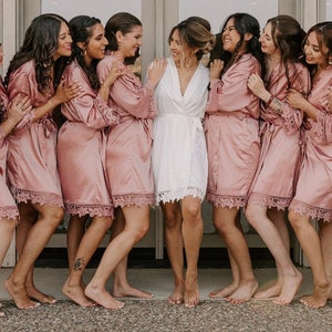 May include: A group of eight people wearing matching pink satin robes with lace trim. The robes are knee-length and have long sleeves. The people are smiling and laughing.