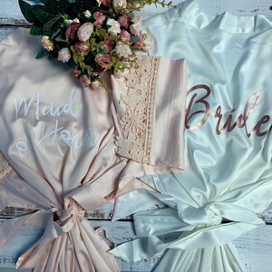 May include: Two satin robes with lace trim. One robe is pink with the text "Maid of Honor" in white. The other robe is white with the text "Bride" in rose gold.