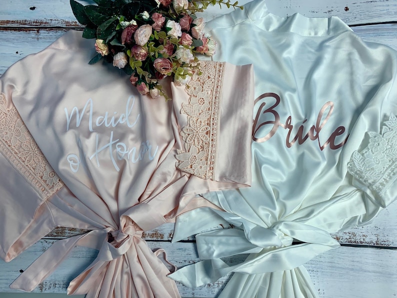 May include: Two satin robes with lace trim. One robe is pink with the text "Maid of Honor" in white. The other robe is white with the text "Bride" in rose gold.