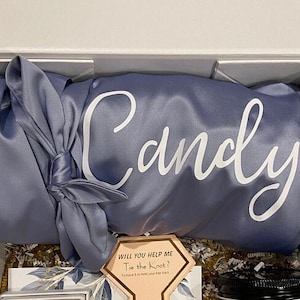 May include: A blue satin robe with the name "Candy" embroidered on it. The robe is folded and tied with a matching blue satin ribbon. A small card with the text "Will you help me tie the knot?" is visible in the foreground.