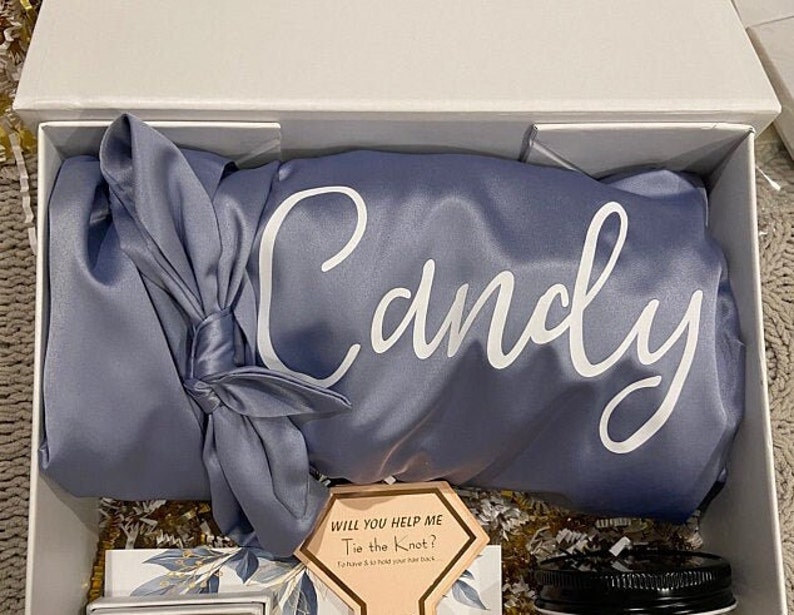 May include: A blue satin robe with the name "Candy" embroidered on it. The robe is folded and tied with a matching blue satin ribbon. A small card with the text "Will you help me tie the knot?" is visible in the foreground.