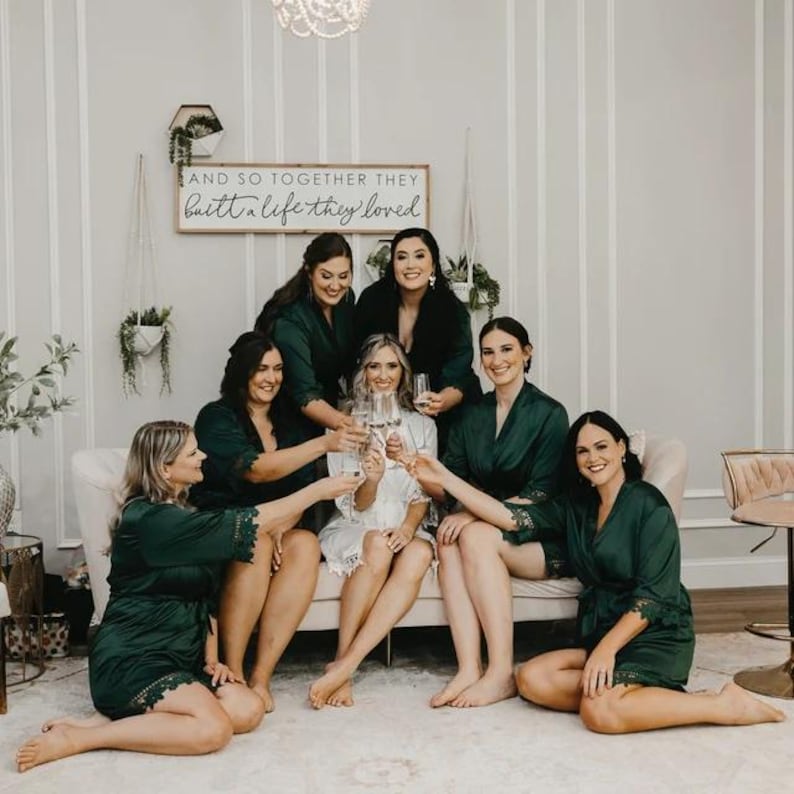 May include: A group of seven women wearing green satin robes with lace trim are gathered together in a room with white walls. They are holding champagne flutes and smiling. A sign above them reads "And so together they built a life they loved."