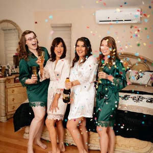 May include: Four women in green and white satin robes celebrate with champagne and confetti. They are smiling and laughing as they raise their glasses.