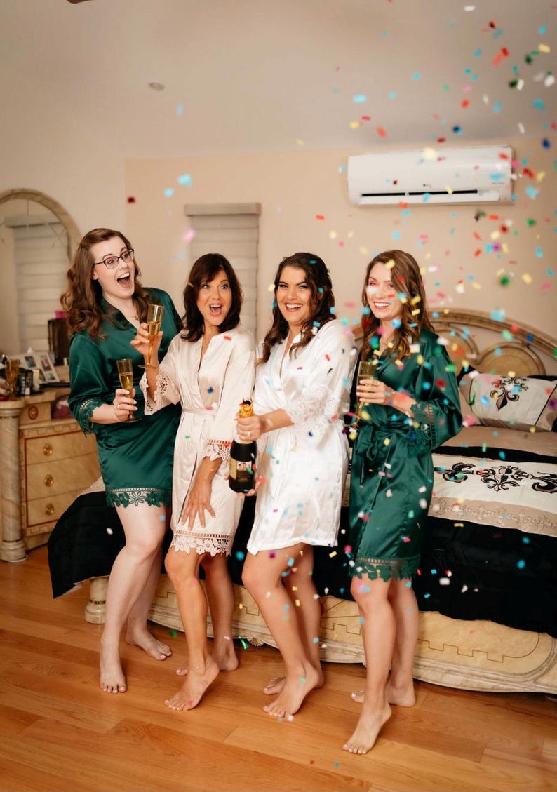 May include: Four women in green and white satin robes celebrate with champagne and confetti. They are smiling and laughing as they raise their glasses.