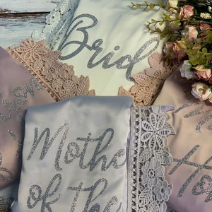 May include: Four satin robes with lace trim and silver glitter lettering. The robes say "Bride", "Maid of Honor", "Mother of the Bride", and "Maid of Honor".