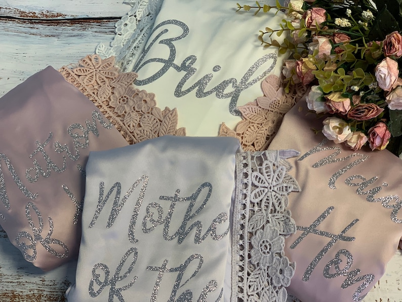 May include: Four satin robes with lace trim and silver glitter lettering. The robes say "Bride", "Maid of Honor", "Mother of the Bride", and "Maid of Honor".