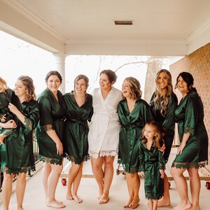 May include: A group of people wearing matching dark emerald green satin robes with lace trim on the sleeves and hems, while one person in the center wears a matching white satin robe with lace trim, all standing together on a porch.