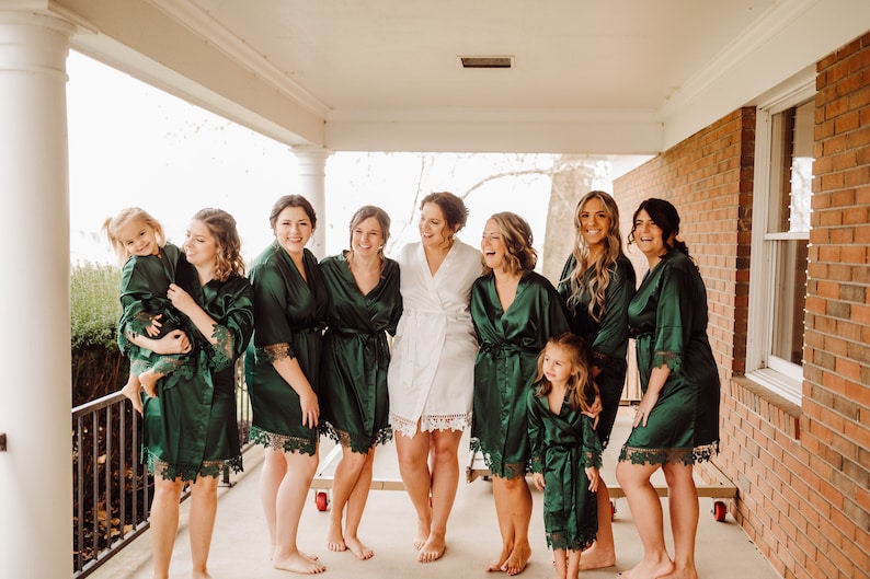 May include: A group of people wearing matching dark emerald green satin robes with lace trim on the sleeves and hems, while one person in the center wears a matching white satin robe with lace trim, all standing together on a porch.