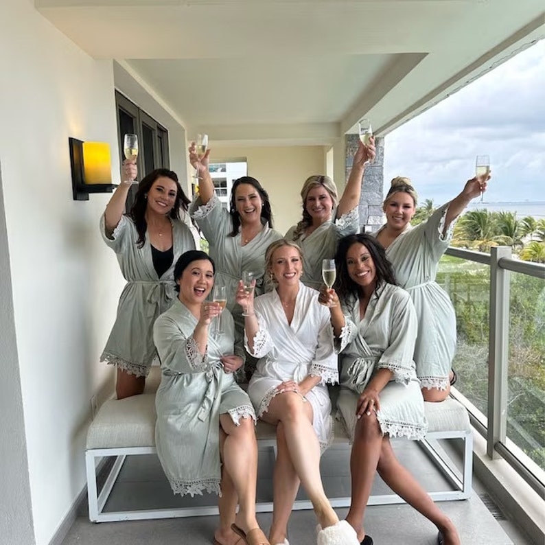 May include: Seven women in light green and white robes are holding champagne flutes and smiling. They are standing on a balcony with a view of the ocean.